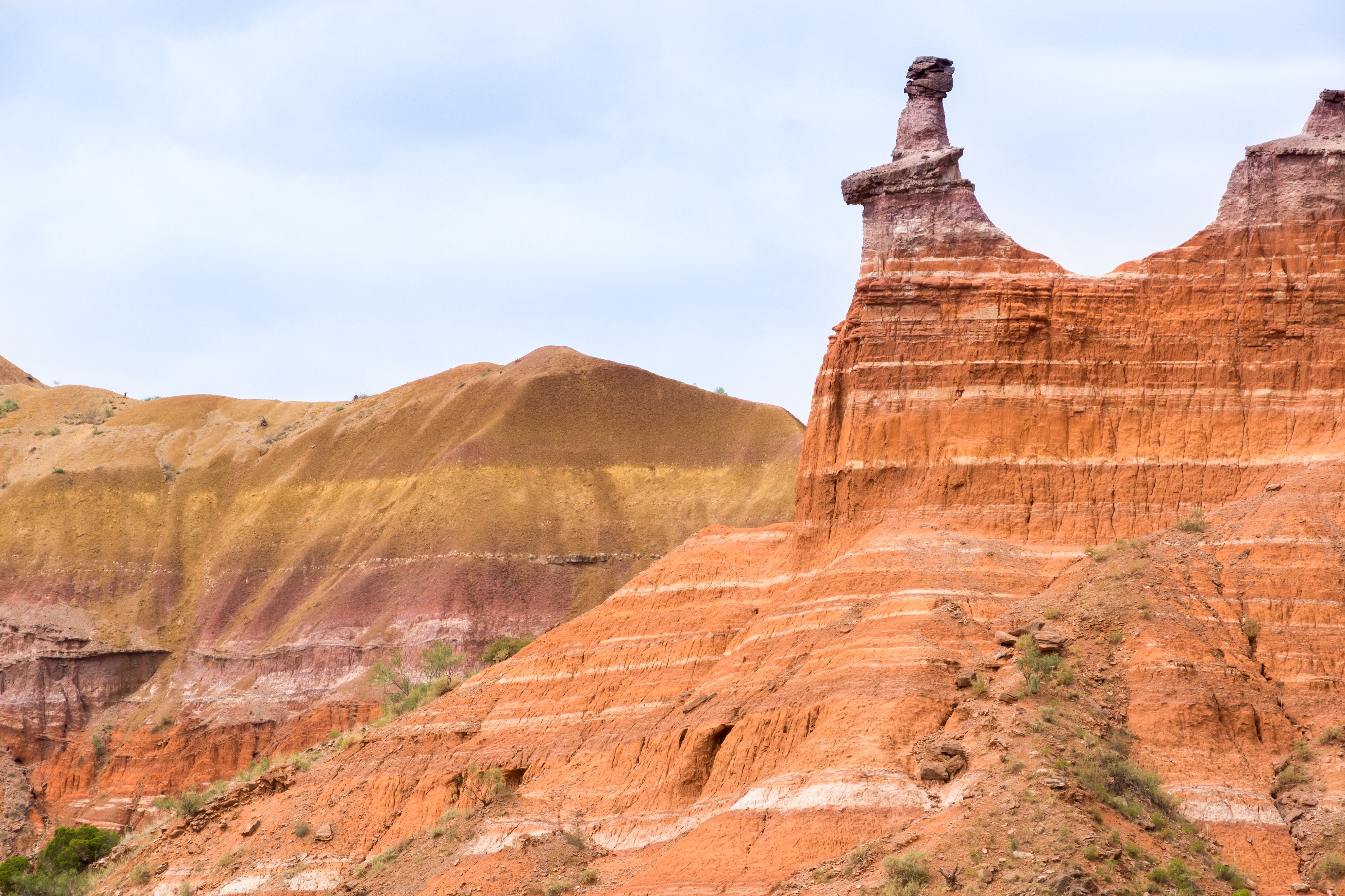 Rock formation stands prominently, resembling a hoodoo, amid layered red and brown cliffs under a clear sky.