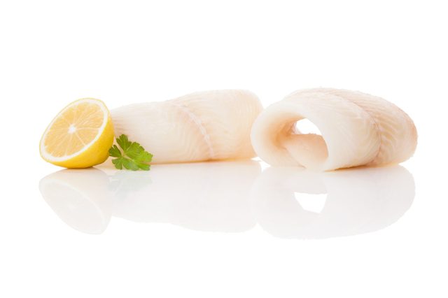 Two raw fish fillets lie on a reflective surface beside a lemon wedge and green parsley sprig, against a white background.