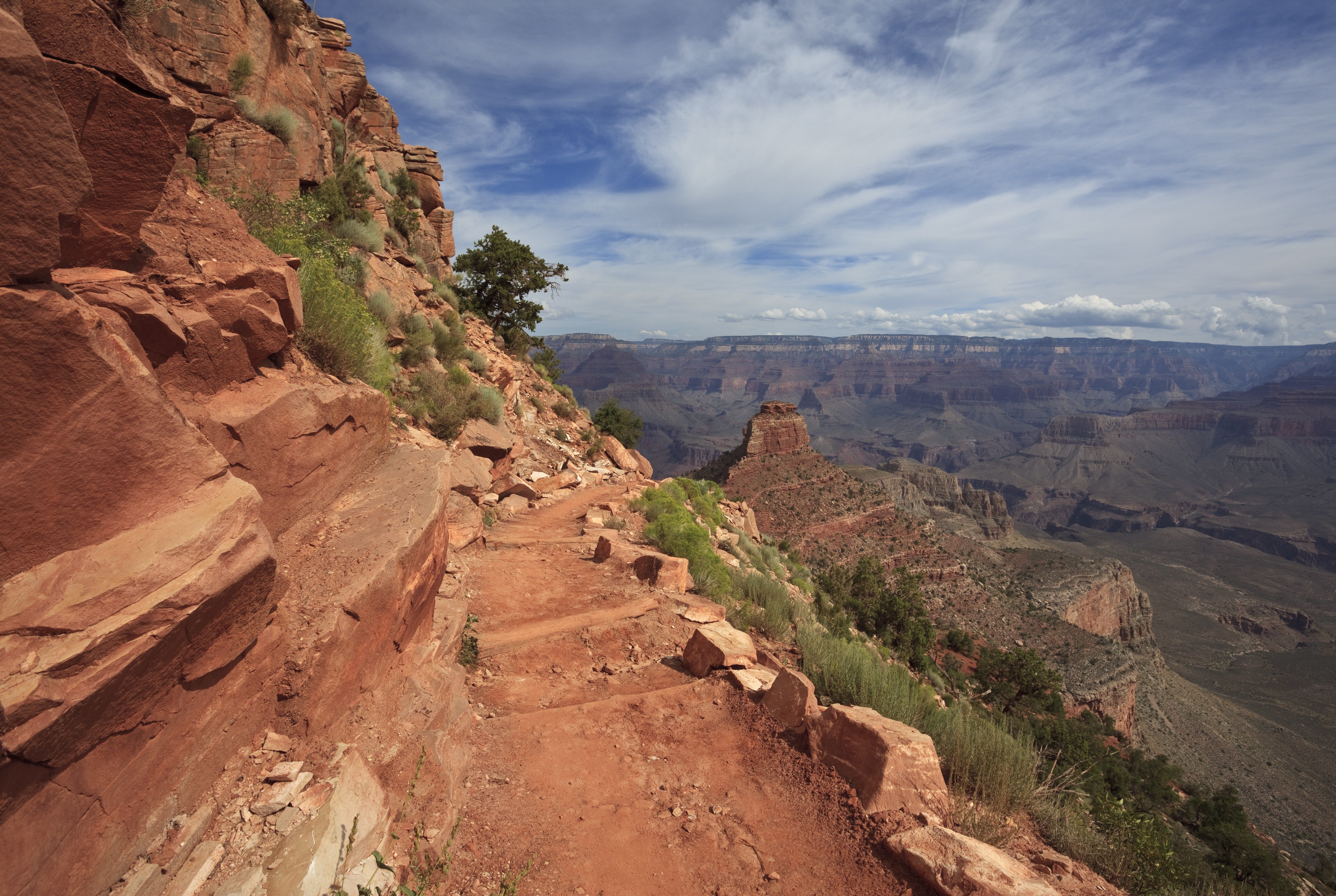 Rugged trail descends along a rocky canyon wall, surrounded by sparse vegetation, against a vast desert landscape and expansive cloudy sky.