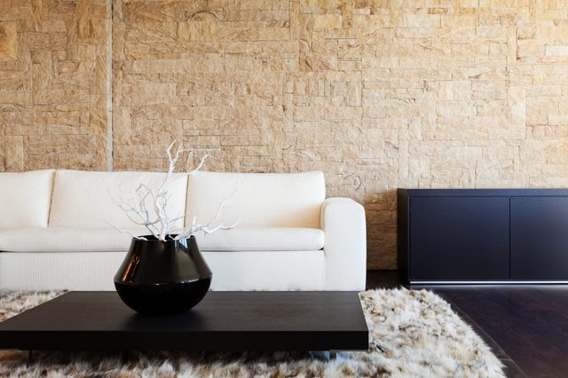 Black vase holds white branches, resting on a modern black table, placed in a living room with a white sofa and textured stone wall.