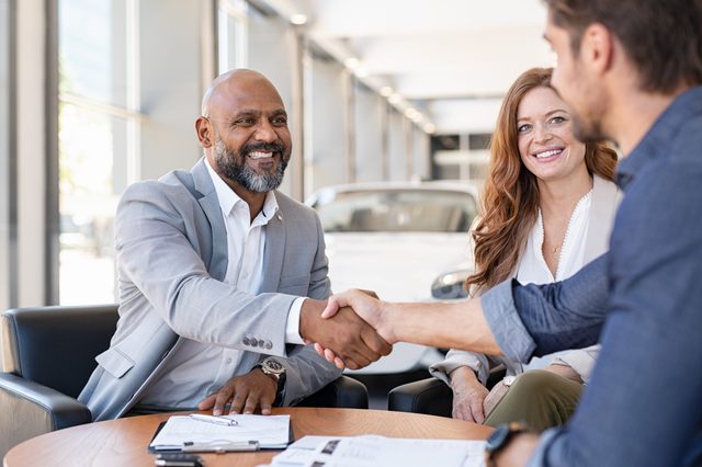 Three people sit in a car dealership; two men shake hands while a woman smiles, surrounded by paperwork and a car in the background.