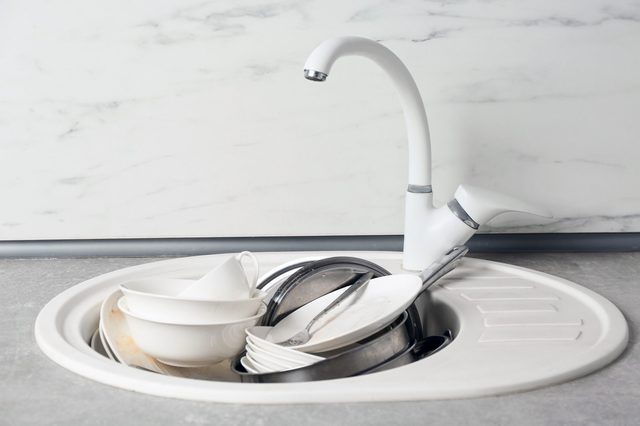 Dishes stacked in a white sink under a curved faucet, set against a light gray countertop and marble-patterned backsplash.