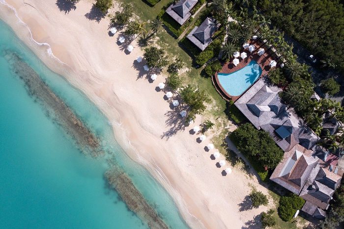 Aerial view of beach umbrellas lining a sandy shore next to turquoise water, adjacent to a resort with a pool surrounded by lush greenery.