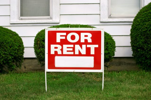 A "FOR RENT" sign stands in front of shrubbery and a white house exterior, on a grassy lawn.