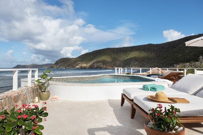 Lounge chairs rest by a pool, overlooking the ocean with lush hills in the background, surrounded by potted plants and a clear blue sky.