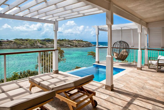 Lounge chairs sit on a shaded terrace overlooking a turquoise sea, with a small pool and hanging chair nearby, surrounded by a scenic coastline.