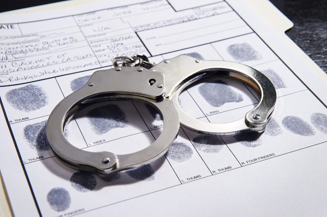 Handcuffs resting on a fingerprint form, surrounded by handwritten personal details and prints, suggesting a law enforcement or criminal background context.