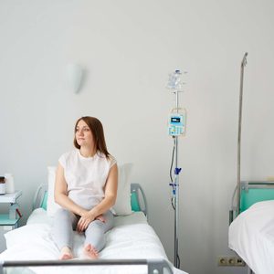 Person sits on hospital bed, calmly looking ahead, with IV stand nearby and another bed beside them in a sterile, minimal hospital room.