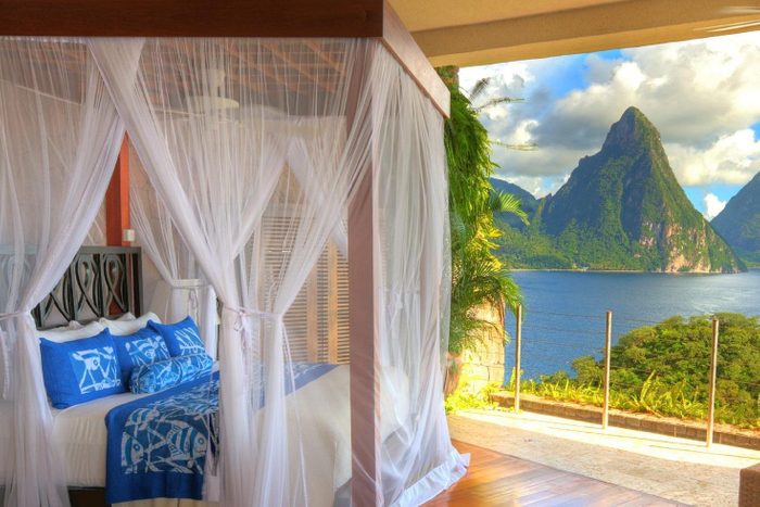 Canopy bed with blue linens sits in an airy room, overlooking lush mountains and a serene blue lake through an open wall.