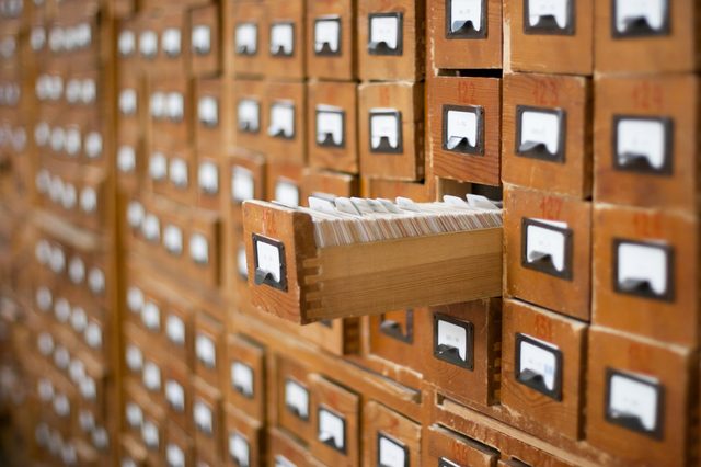 Wooden library card catalog drawer extends outward, revealing index cards, amidst rows of identical drawers with labeled handles.