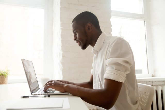 Person typing on a laptop inside a bright room, with pen and plant on the table, situated near large windows.