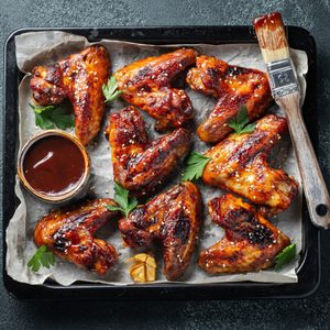 Chicken wings glazed in barbecue sauce rest on a parchment-lined tray with a basting brush and dip, garnished with parsley and garlic.