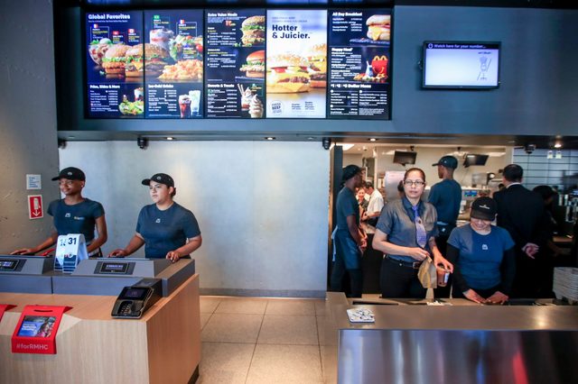 Fast-food employees working at counters under bright menu boards in a restaurant, with some people preparing food in the background.