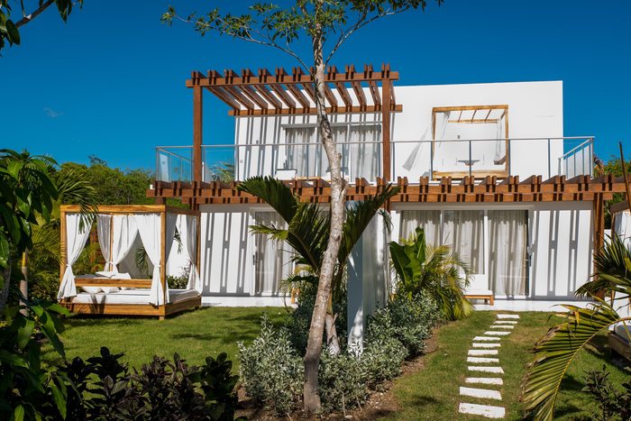 Modern villa with wooden pergolas and draped cabana on lush green lawn, surrounded by tropical plants under a clear blue sky.