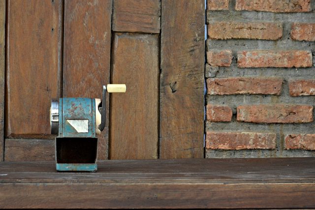 An old blue pencil sharpener with a manual handle sits on a wooden table, backed by a rustic wooden and brick wall.