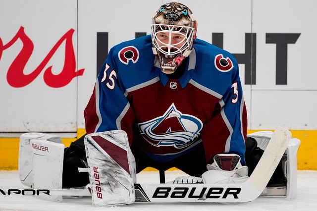 Hockey goalie crouches on ice wearing maroon and blue jersey, with "35" visible, in front of a partially visible advertisement.