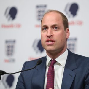 A man speaks into a microphone, wearing a blue suit and red tie, in front of a backdrop with logos and 