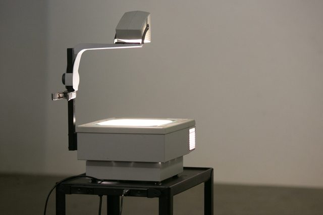 An overhead projector illuminates, casting light onto a blank wall, in a dimly lit room, with cables trailing onto a small table.