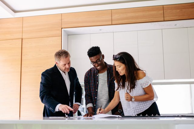Three people stand at a kitchen counter, discussing and signing paperwork, with wooden cabinets and a modern aesthetic in the background.