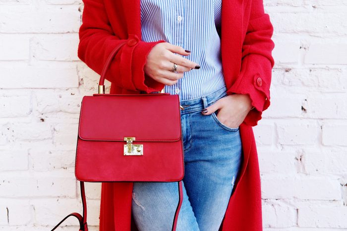 Closeup of red small bag in hand of woman. Fall spring fashion outfit red coat and trendy blue jeans
