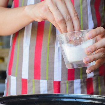 Hands sprinkle salt from a glass cup over a pot, wearing a colorful striped apron in a kitchen setting.