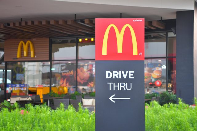 A McDonald's sign directs cars to the drive-thru, positioned near a restaurant with visible windows and surrounding greenery.