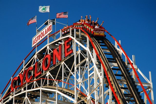 Roller coaster descends steep track; passengers raise hands. Surrounding flags and blue sky. Signs read "CYCLONE" and "ASTROLAND."