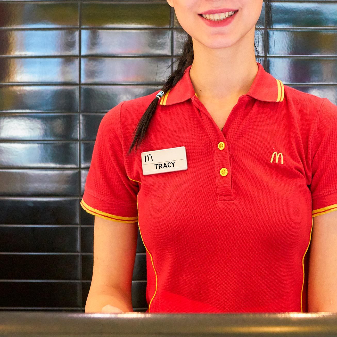 Employee stands smiling in front of a black-tiled wall, wearing a red McDonald's uniform with "Tracy" name tag.