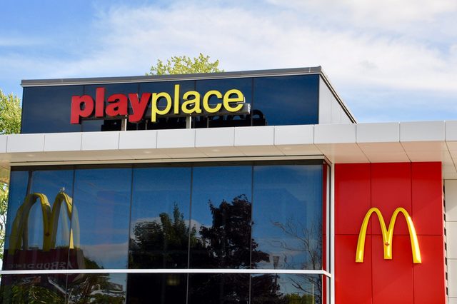 Building with "playplace" sign, featuring yellow and red logo, reflecting trees and sky in glass windows, surrounded by greenery.
