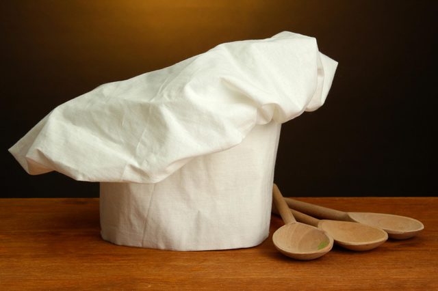 Chef's hat rests beside three wooden spoons on a wooden surface, set against a dark background.