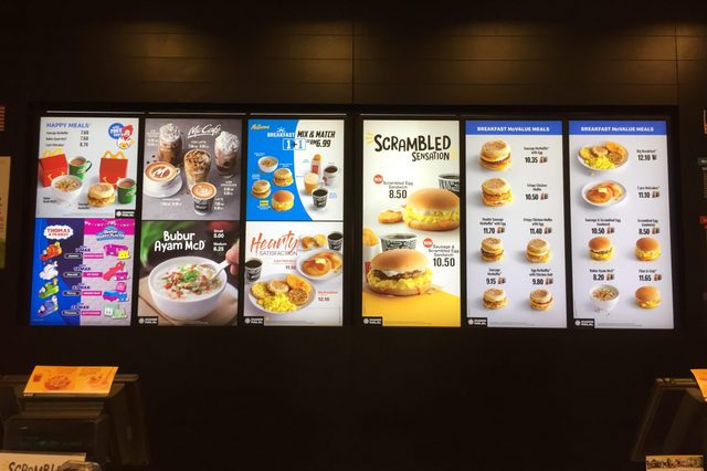Digital menu displays various breakfast items, including sandwiches, muffins, and meals, alongside prices in a fast-food restaurant setting.