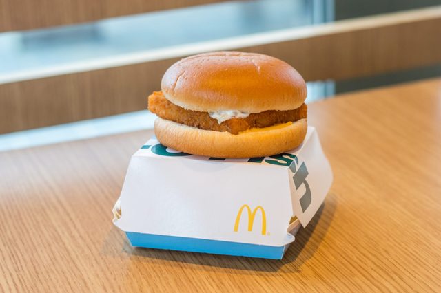 A fish sandwich rests on a branded box, placed on a wooden table in a fast-food restaurant setting.
