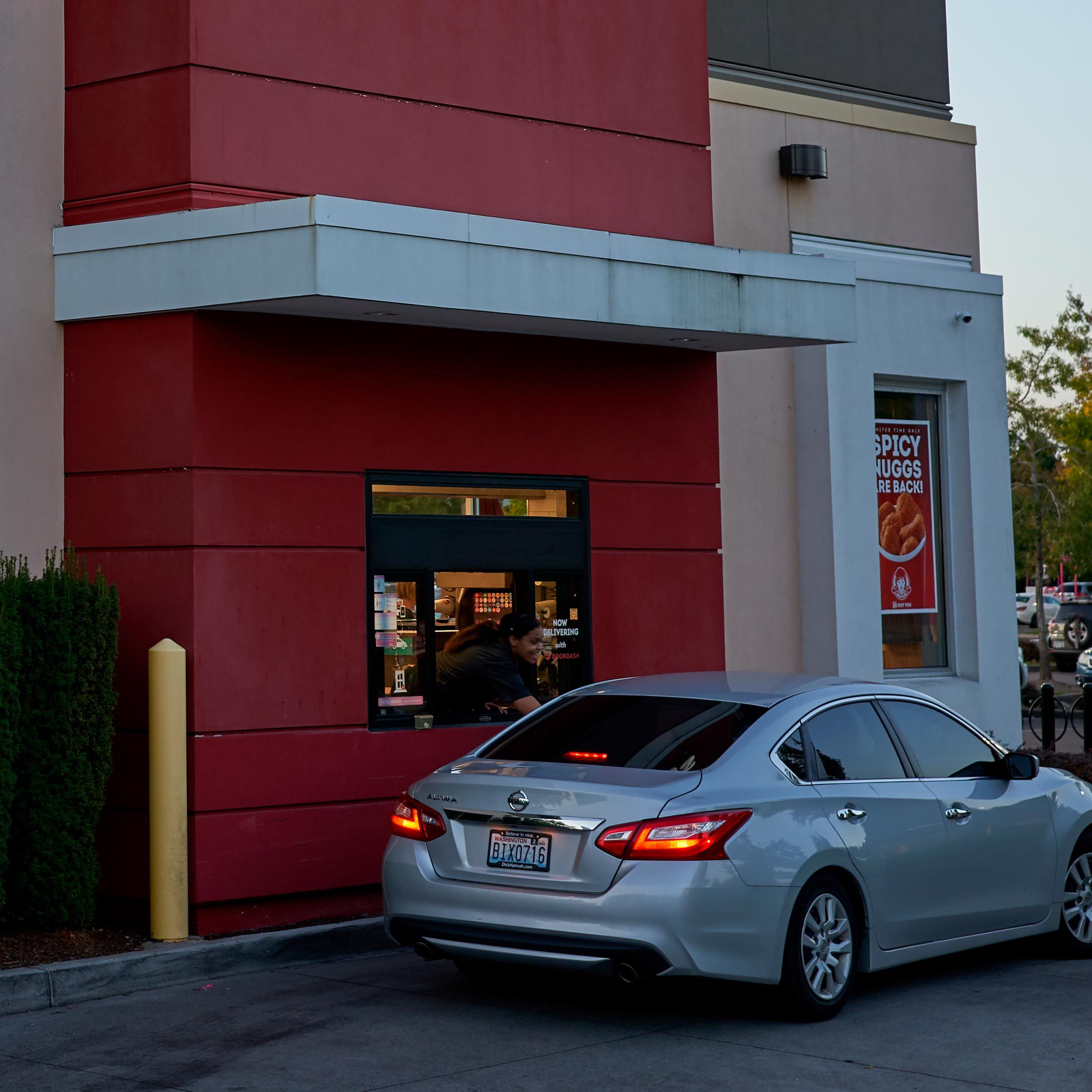 Car waits at drive-thru window; employee leans out, handing item; red restaurant building with "Spicy Nuggs Are Back!" poster visible.