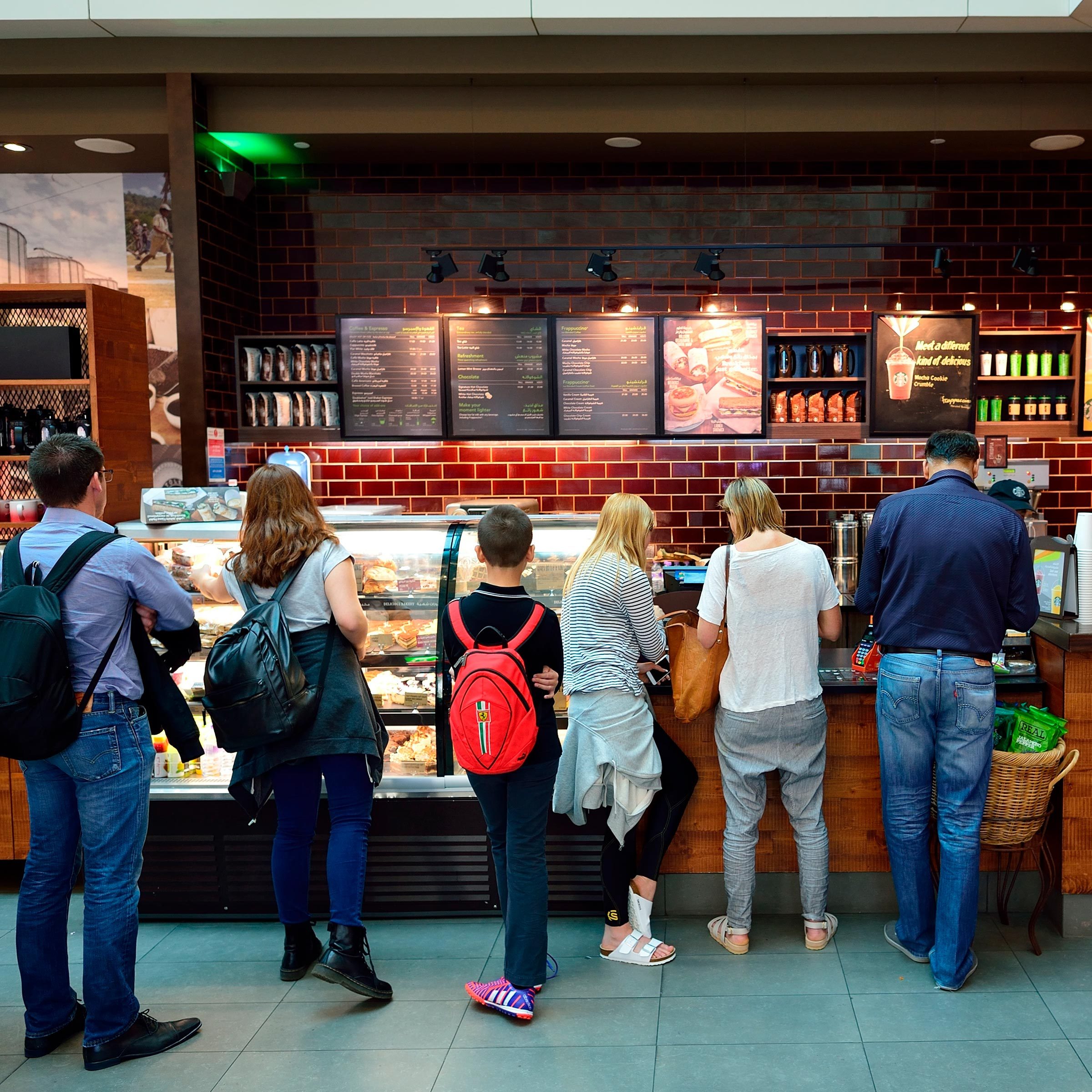 People wait in line, ordering at a coffee shop counter; menus and display cases with food items fill the bustling, brick-walled cafe environment.