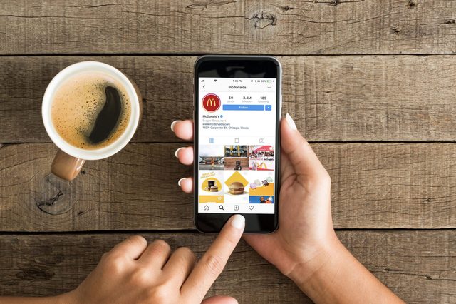 Hand holding smartphone browsing Instagram profile; wooden table surface; coffee cup nearby.