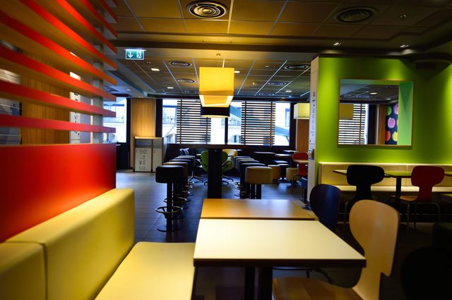Chairs and tables arranged neatly in a colorful, modern restaurant with bright lighting, red-and-green decor, and large windows with wooden slats.