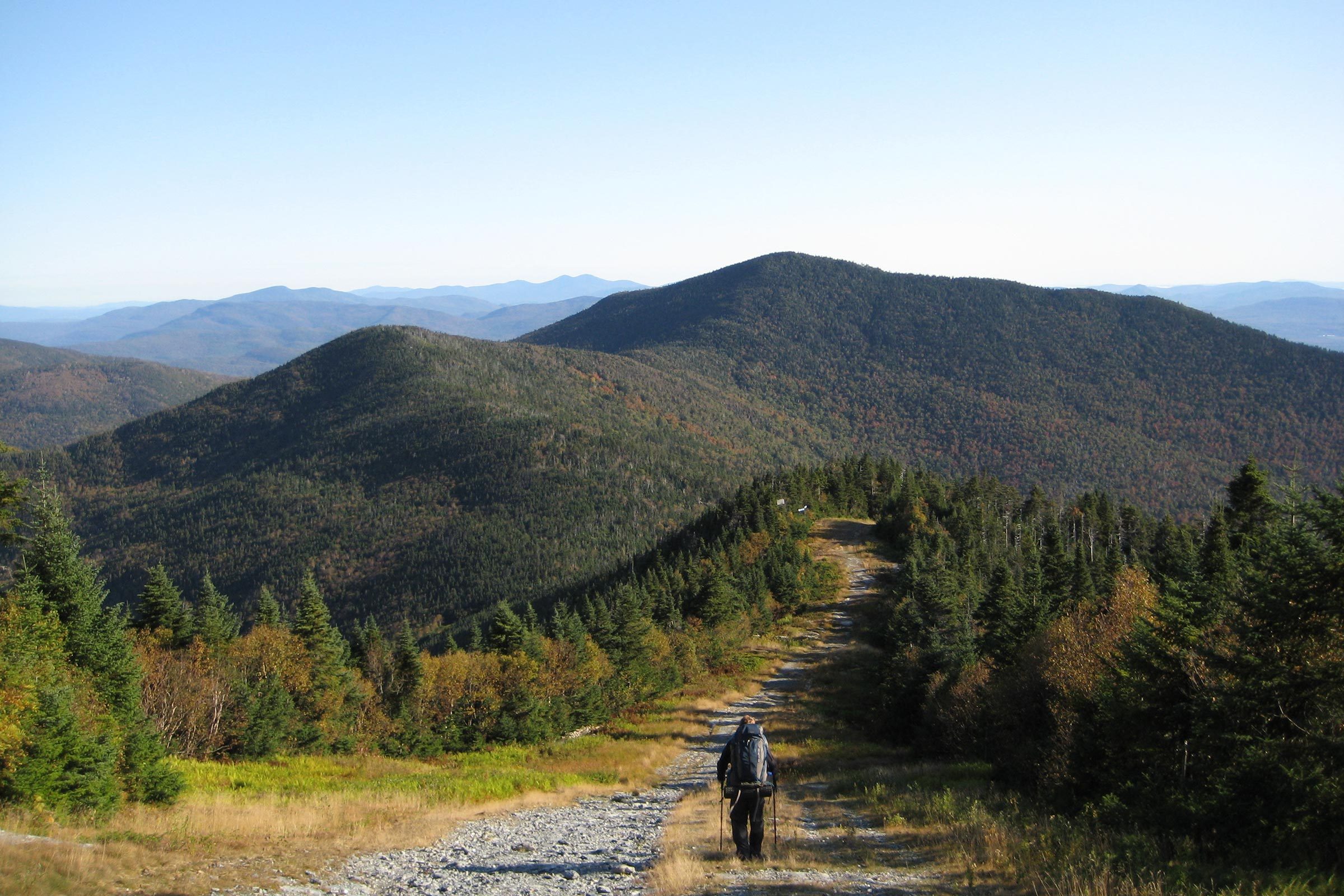A hiker walks along a mountain trail surrounded by dense forest, with distant rolling hills under a clear blue sky.