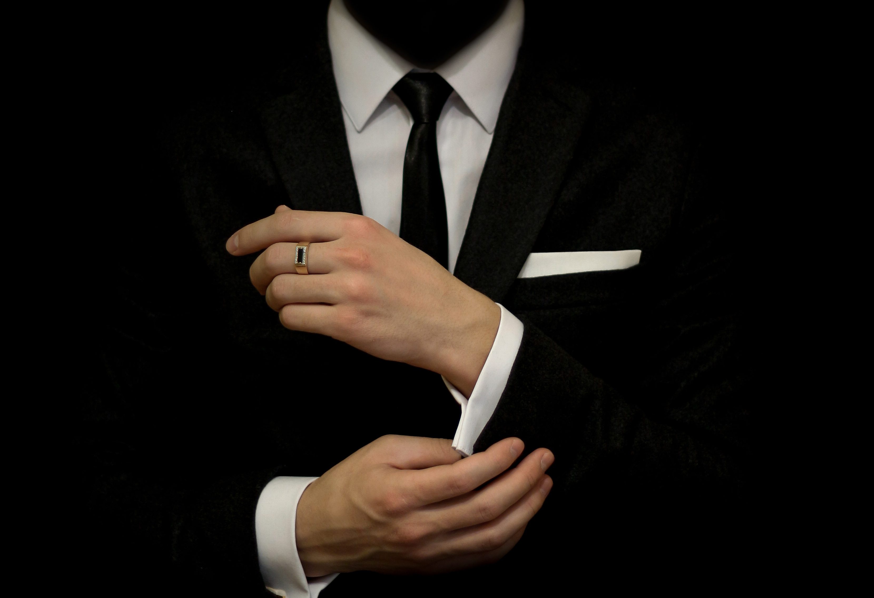 Person adjusts cuff of a black suit, wearing a tie and ring, in a dark setting.