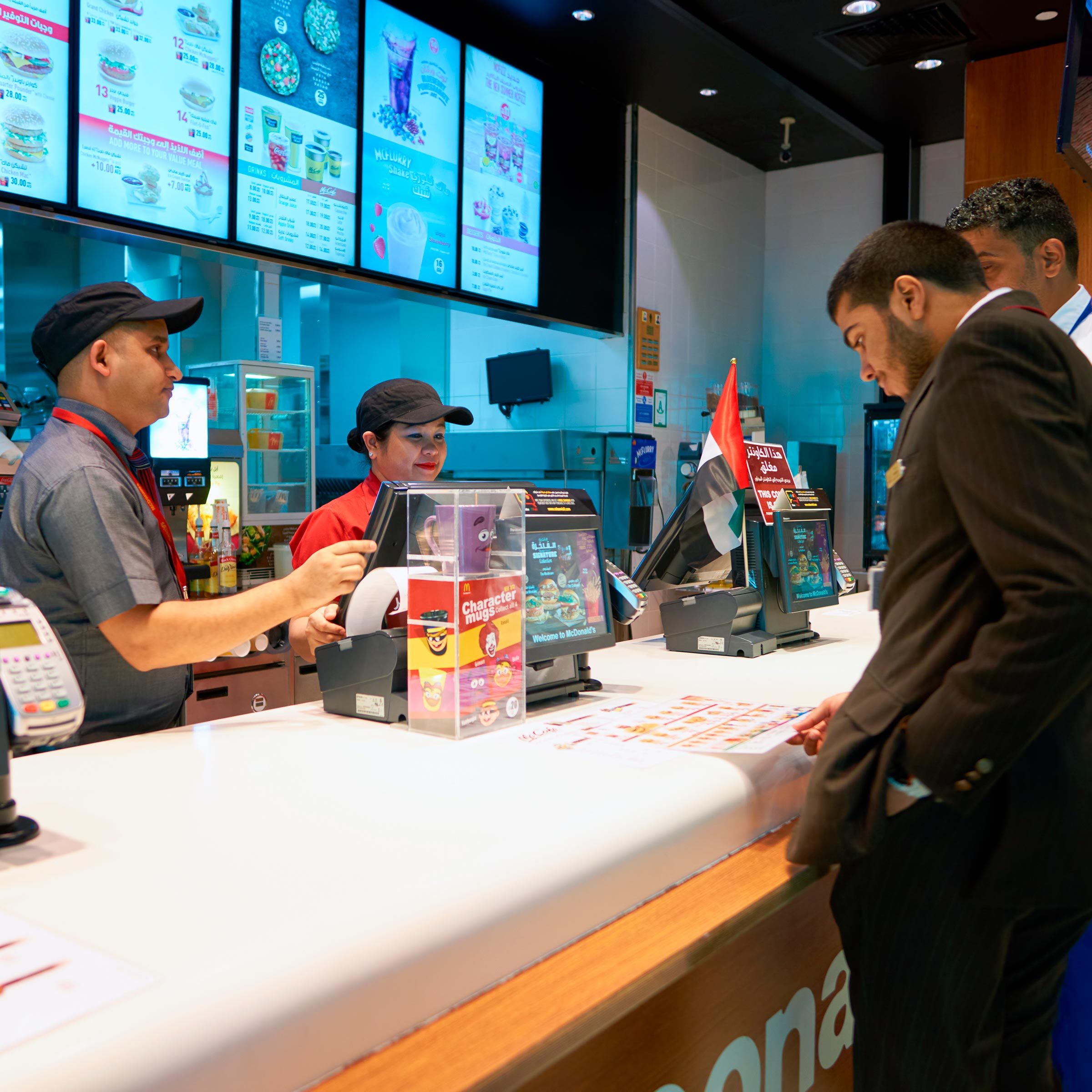 A customer ordering food at a fast-food counter, with employees assisting, in a brightly lit restaurant displaying menus on digital screens.