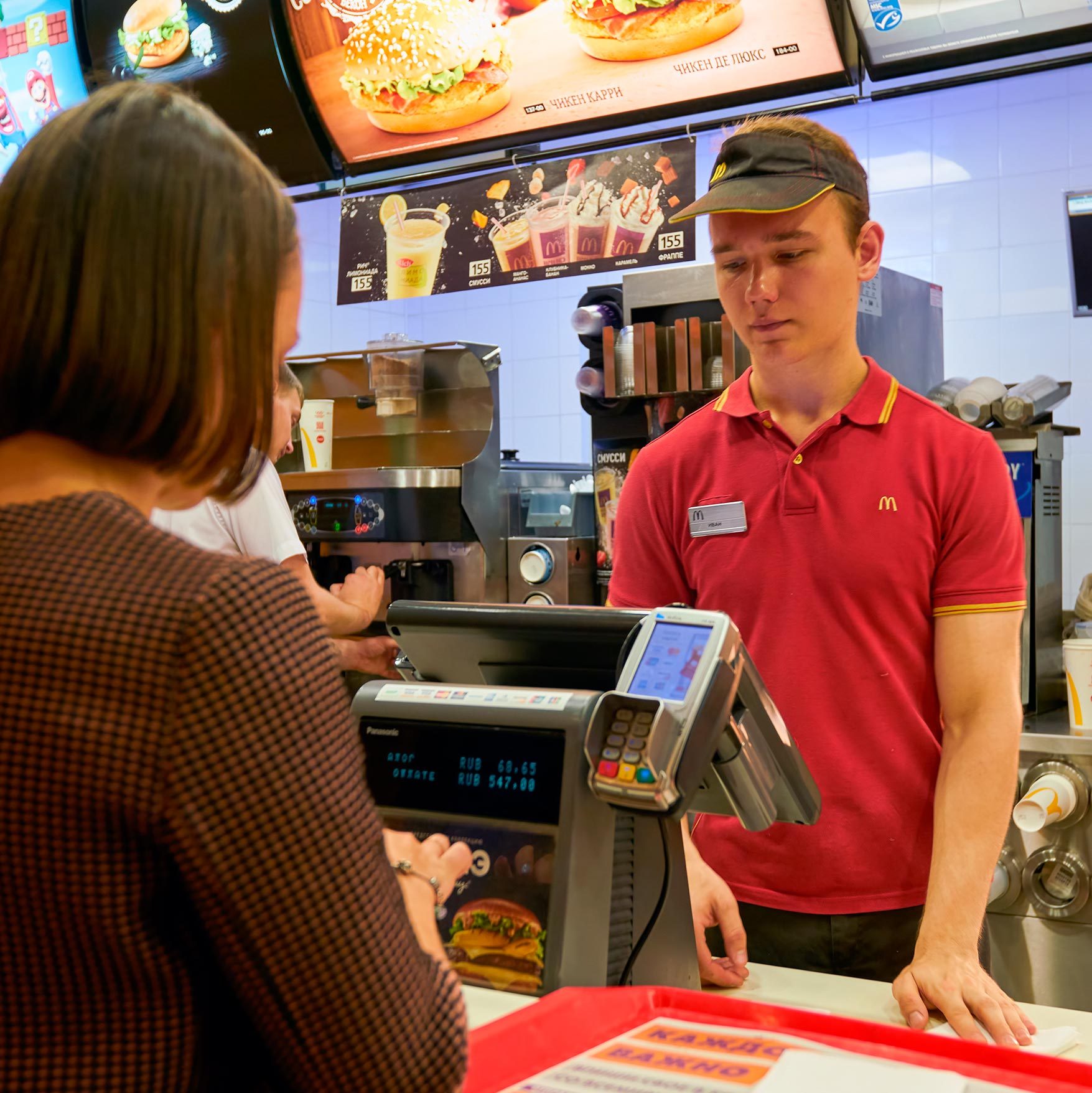 Cashier stands at counter, assisting customer in fast-food restaurant; menu boards display food options above.