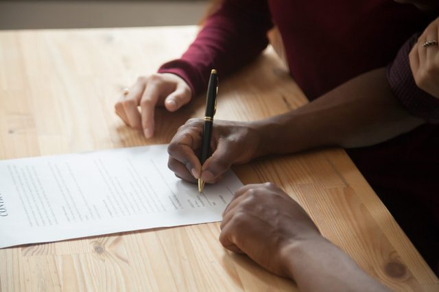 Hand signing document on wooden table, accompanied by another hand pointing, indicating collaboration or guidance in the process.