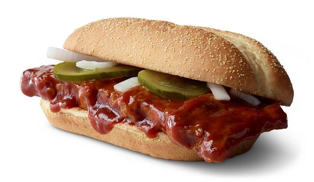 A sandwich holds a barbecue-sauce-coated rib patty with pickle slices and onions on a hoagie roll, isolated against a white background.