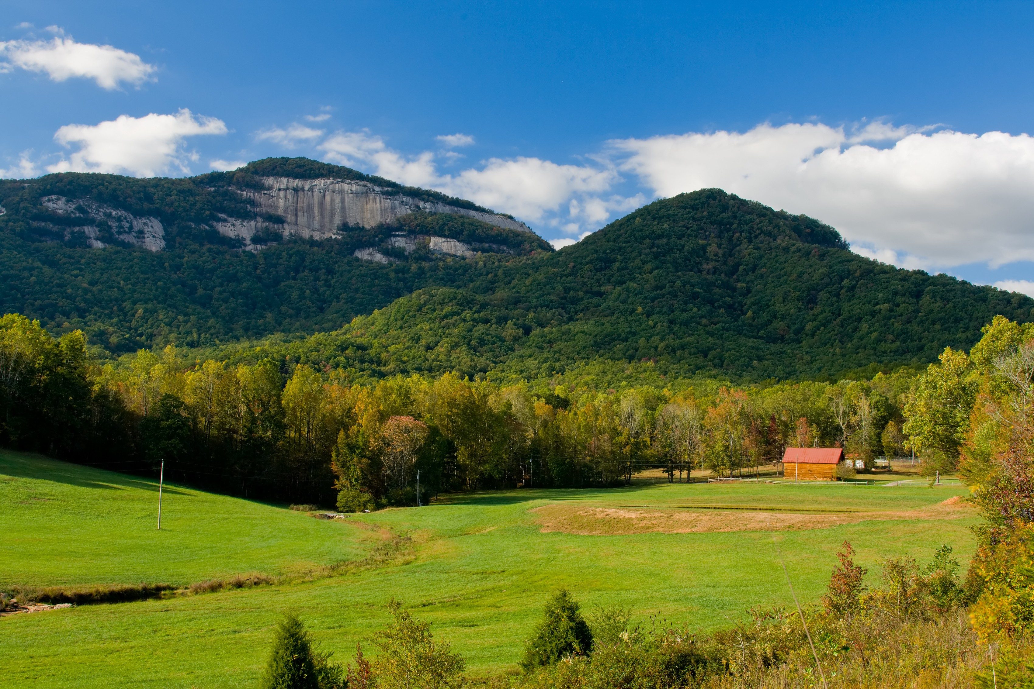 A red-roofed barn sits amidst green fields with a forest in the background, framed by lush, tree-covered mountains under a blue, partly cloudy sky.