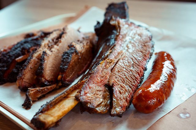Barbecue meat rests on parchment paper, featuring slices of brisket, a large rib, and a sausage, with a wooden table underneath.