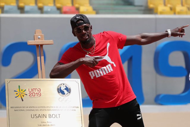 Individual poses energetically, pointing upward, in front of a commemorative plaque. The background features stadium seating and large blue numbers.