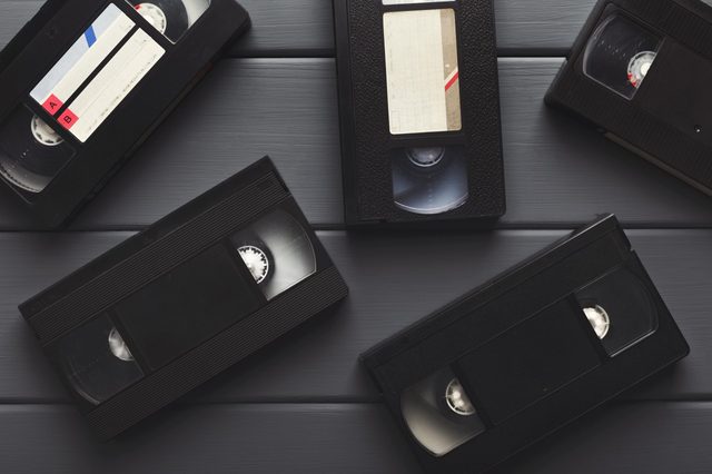 VHS tapes lay scattered on a dark wooden surface, some with blank labels, suggesting retro media storage in a simple arrangement.