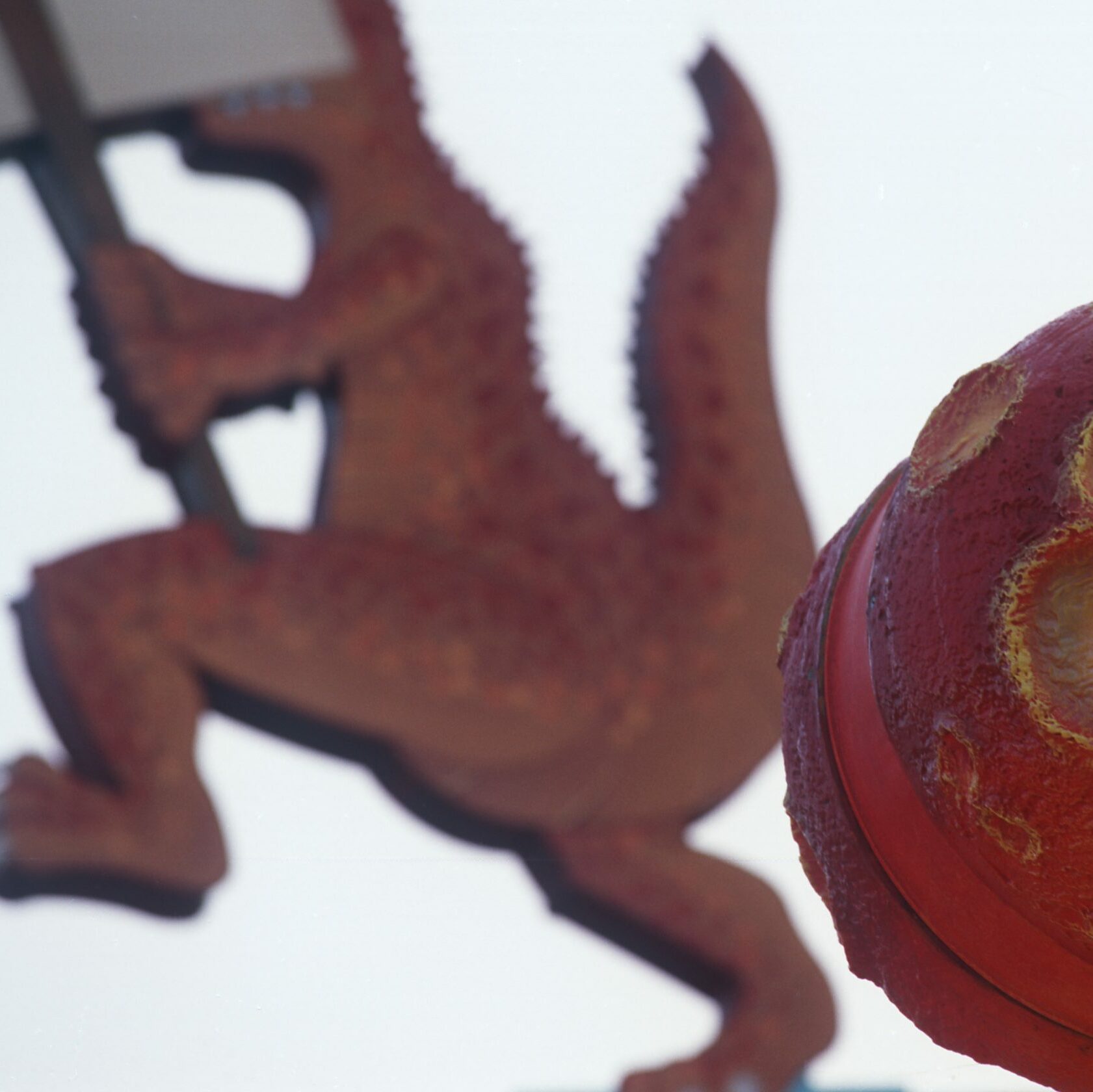Red textured sphere close-up, resembling a moon, with a blurry dinosaur figure holding a sign in the background against a clear sky.