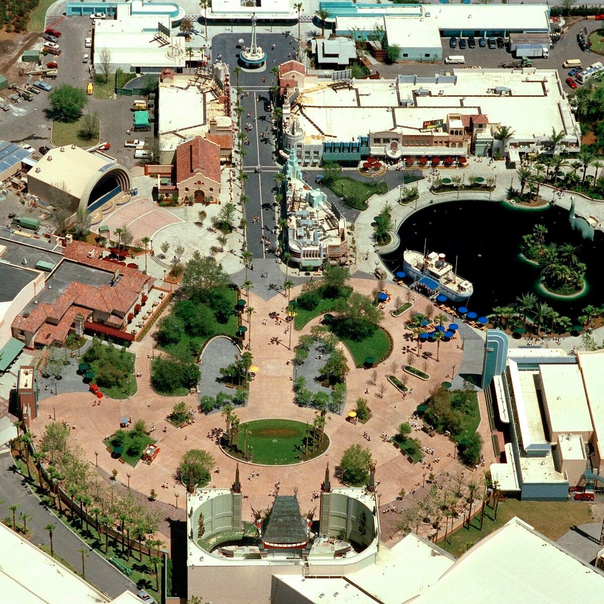 Aerial view of theme park plaza with pathways, greenery, and buildings surrounding a pond with a boat. Visitors walk and gather in the area.