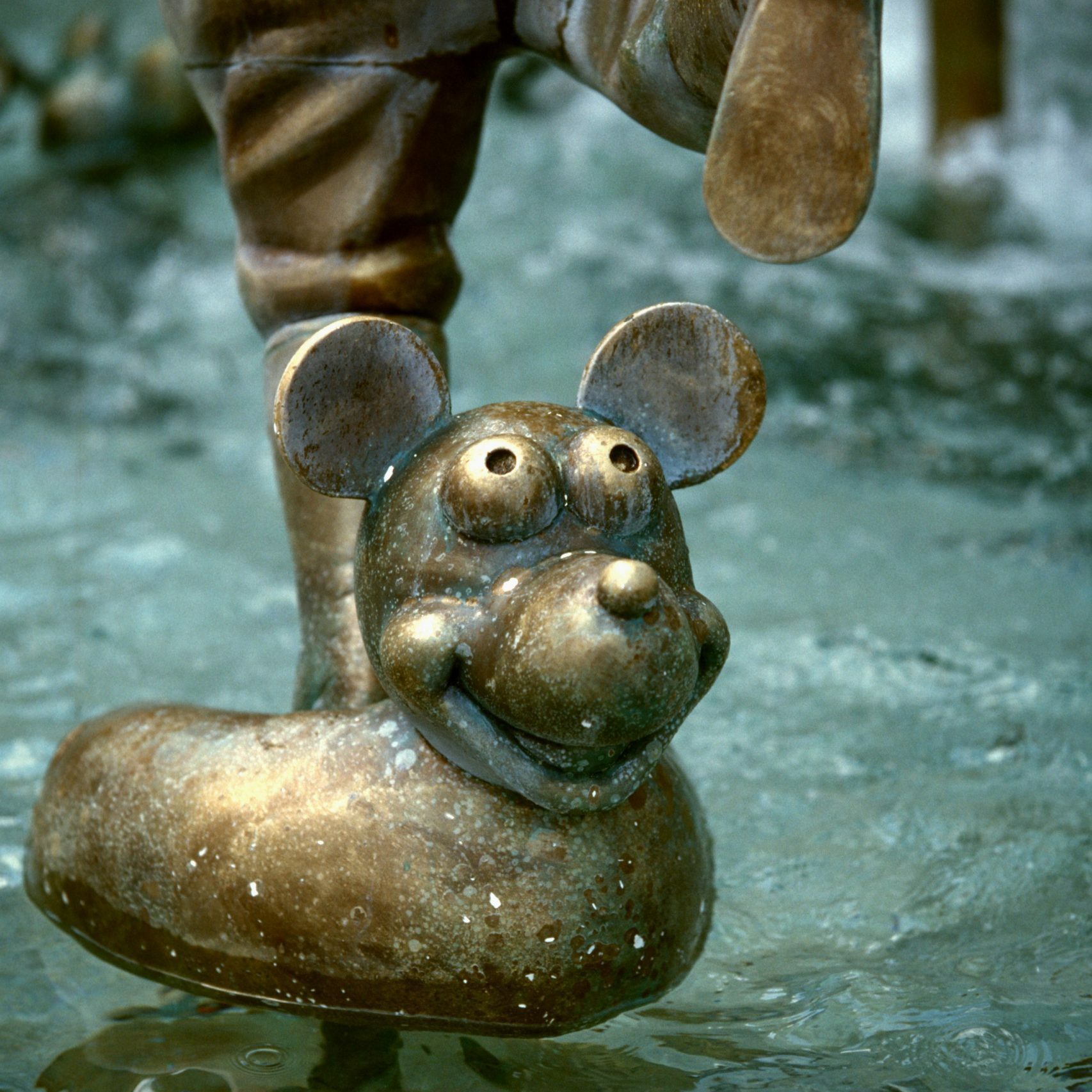 Bronze mouse sculpture smiles, resting near a playful fountain's water, evoking whimsy and charm in a public space.
