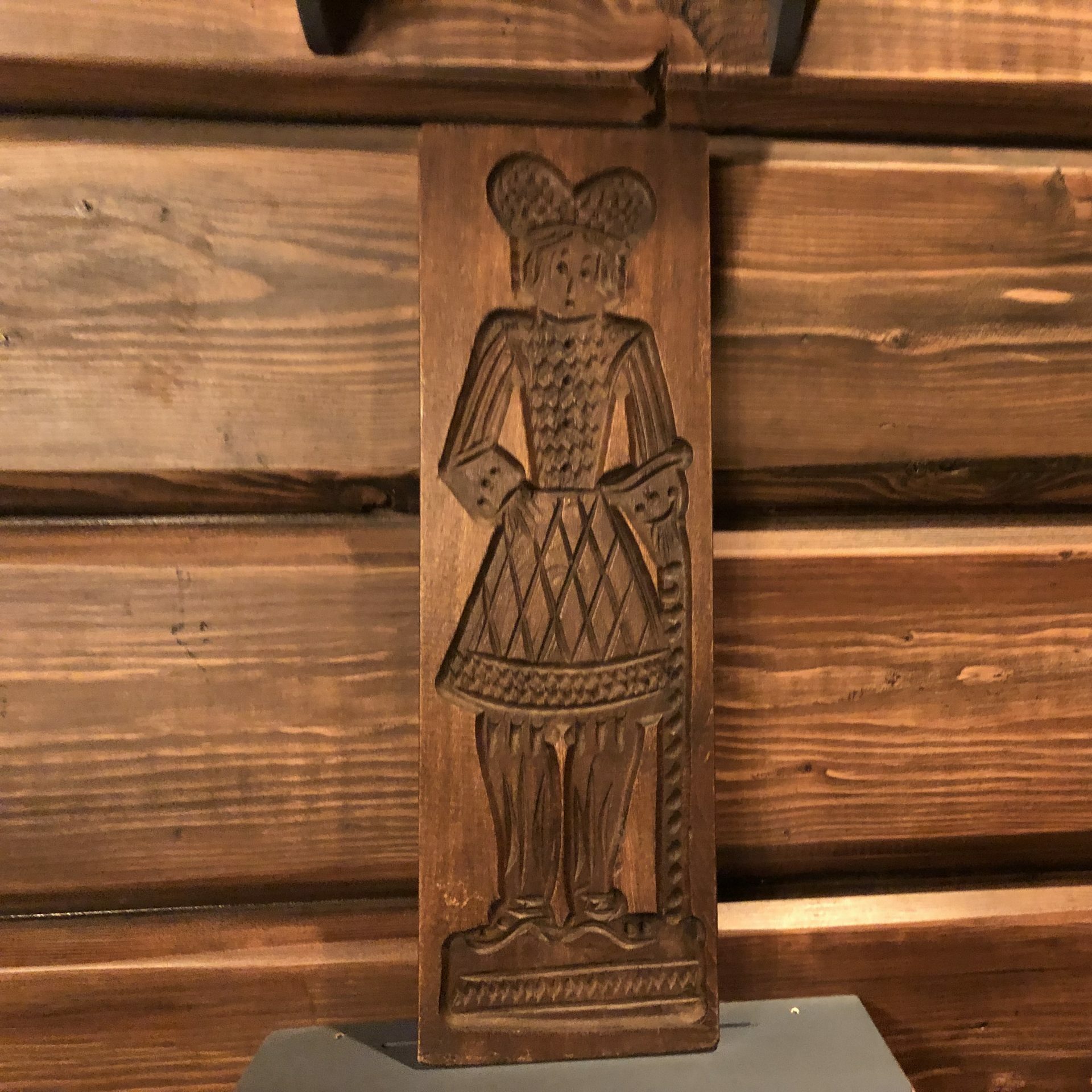 Carved wooden panel depicts a figure in traditional attire, standing upright. The background consists of horizontally aligned wooden planks.